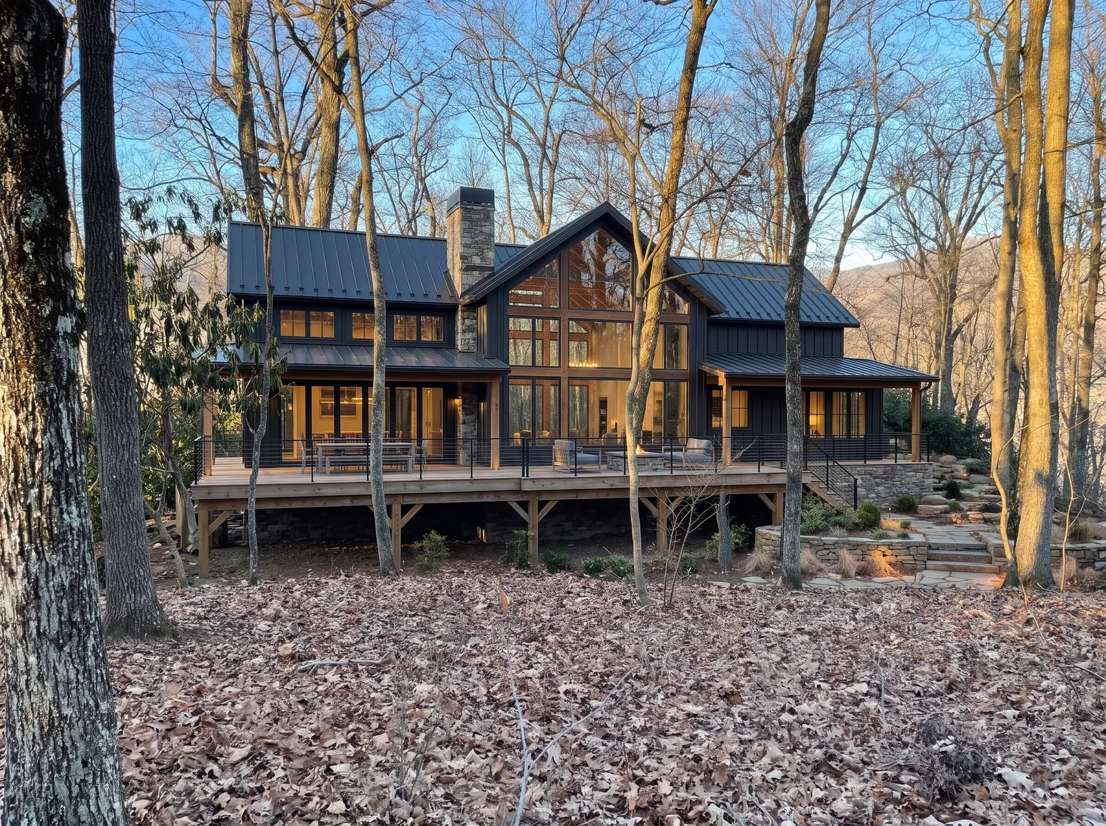 Concept rendering: a two-level dark modern mountain home with a metal roof, stone chimney and a wall of glass among oaks at golden hour, with a large deck on timber posts over the slope, a dining table, lounge seating and a fire table