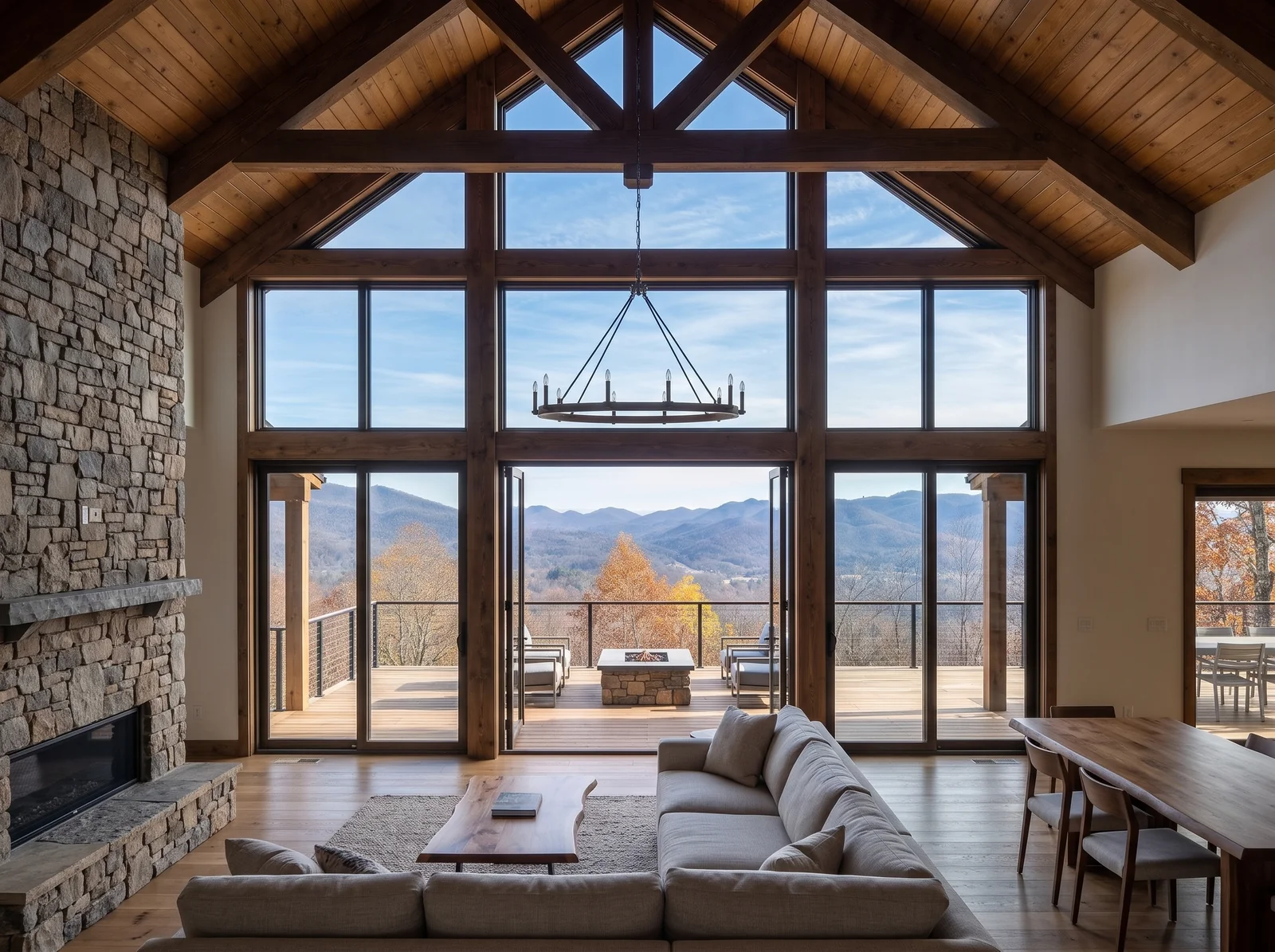 Concept rendering: the great room inside the glass gable, with a stone fireplace, a vaulted wood ceiling with a timber truss, and floor-to-peak glass looking out over the deck and fire table to layered ridgelines
